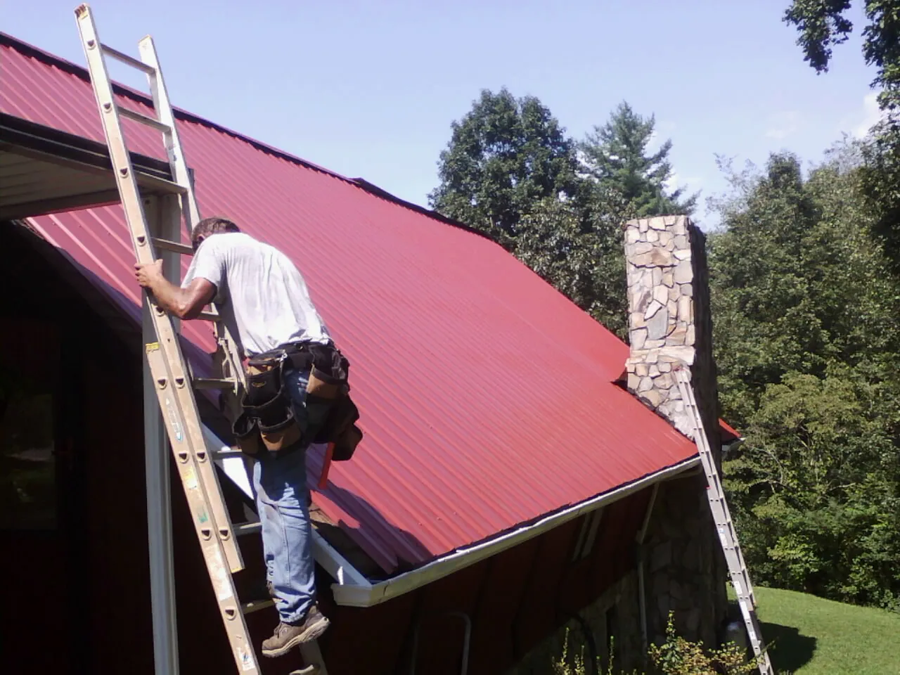 Roofer on a ladder beside a red metal roof and stone chimney — Soffit & Fascia Repair by Roofing Brothers in Wilmette